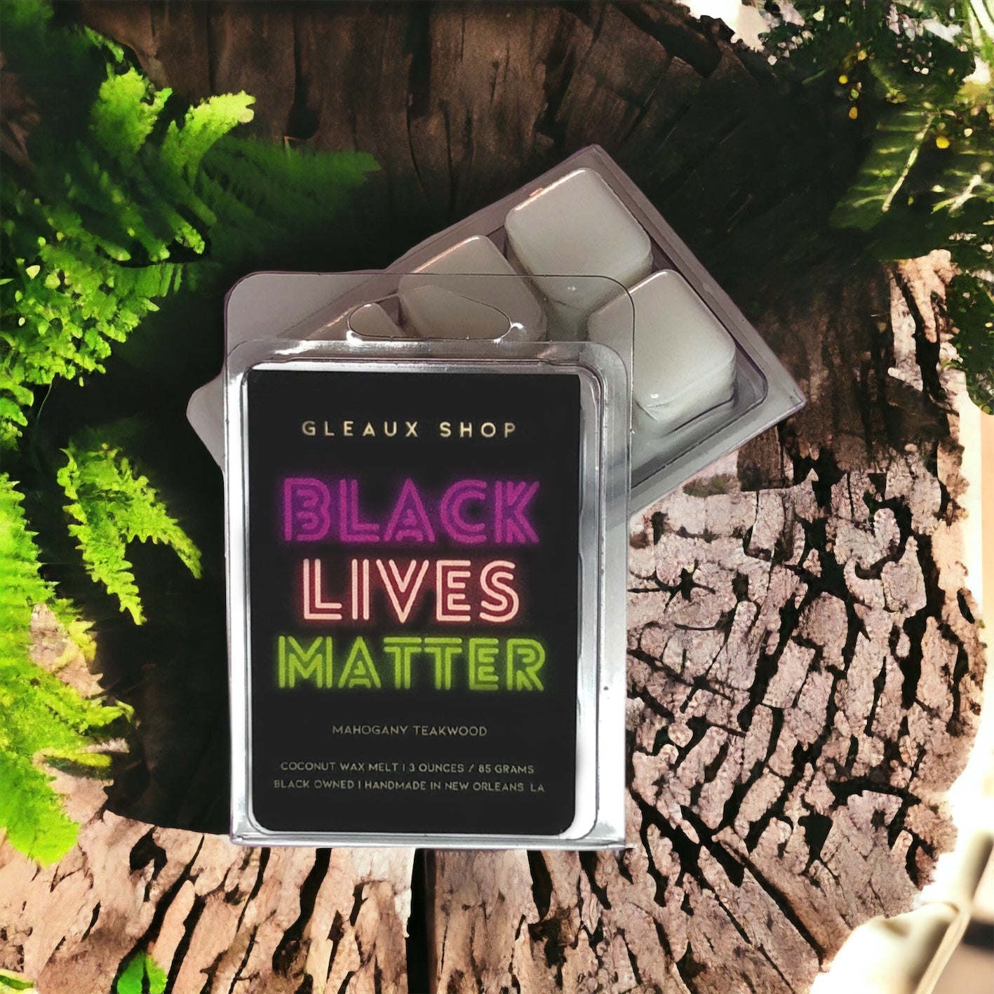 A package of 'Black Lives Matter' scented wax melts by Gleaux Shop, with the packaging open and revealing the wax melt discs inside. The packaging has a black background with pink and green text, and is placed on a rustic wooden surface with greenery in the background.