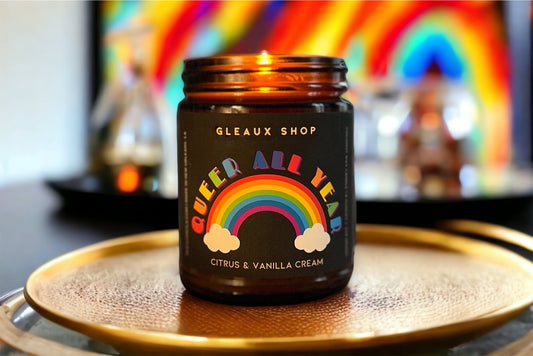 Queer All Year citrus and sandalwood scented coconut wax candle by Gleaux Shop in an amber jar with a black label featuring a colorful rainbow design. The jar is placed on a round, silver tray, and there is a multicolored background.